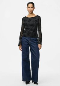 Maglietta nera a maniche lunghe con sovrapposizione di motivo floreale, abbinata a jeans in denim a gamba larga. Il look è completato da tacchi neri.