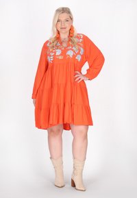 Orangenes gestuftes Kleid mit floraler Bestickung, langen Ärmeln und V-Ausschnitt. Getragen mit beigen Cowboy-Stiefeln. Glatte Textur, lässiges Design.