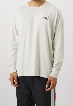 Mężczyzna w białej koszuli z długim rękawem z małym czerwono-czarnym logo Under Armour i czarnych spodniach z logo obok kieszeni.