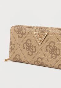 Béžová peňaženka s diamantovým vzorom a vyrazenými písmenami. Obsahuje zlaté trojuholníkové logo a zapínanie na zips. Hladká textúra.