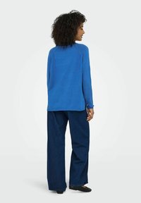 Blå långärmad topp med rundad halsringning och raglanärmar, tillsammans med lösa mörka denimbyxor och platta svarta skor.