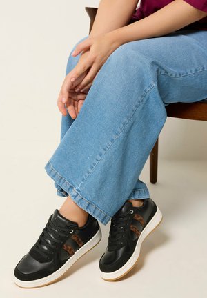 Personne assise sur une chaise en bois portant un pantalon en denim bleu clair et des baskets noires avec des rayures imprimées léopard marron.