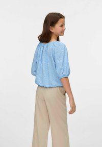 Lichtblauwe blouse met wit bloemenpatroon, geplooide mouwen en elastische zoom. Gepaard met beige, wijde broek.