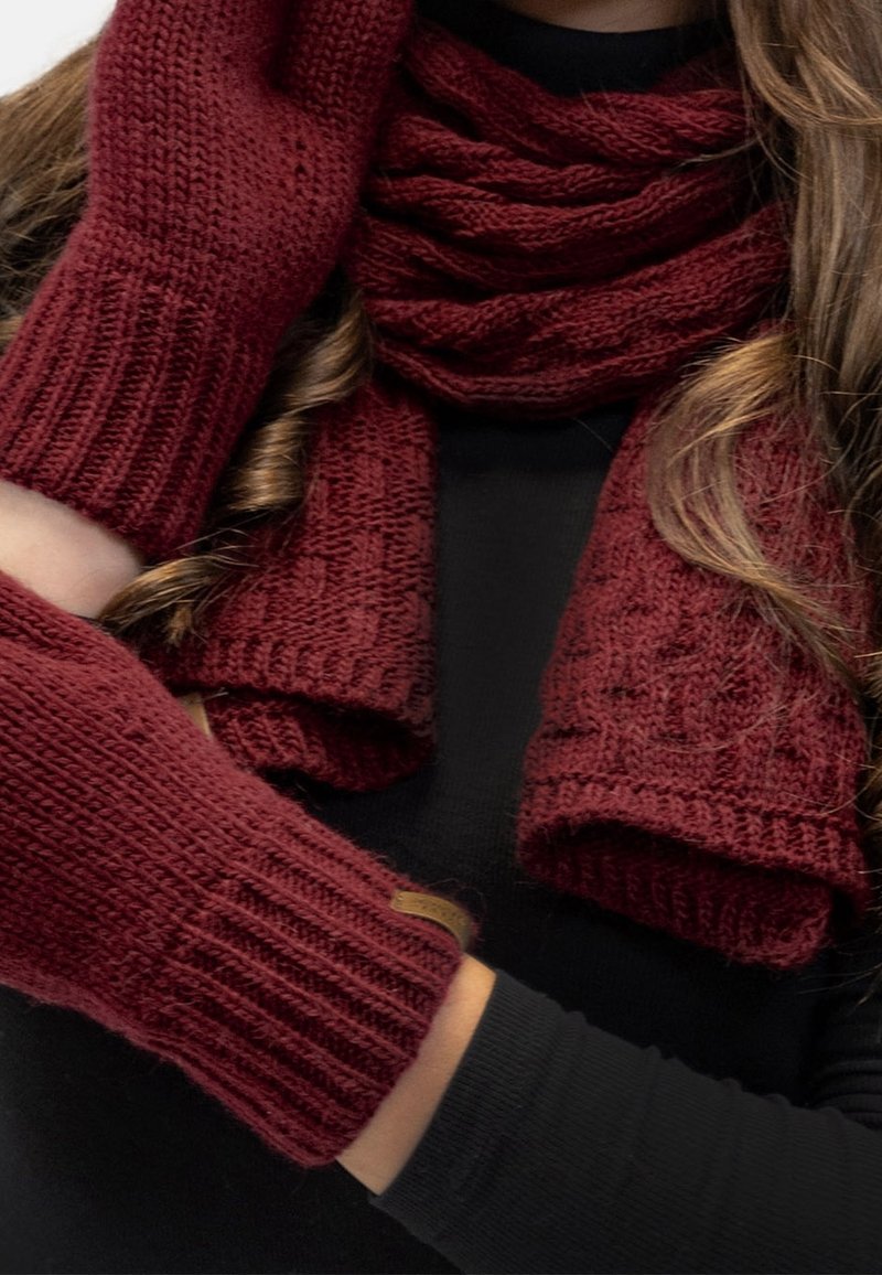 Femme portant une écharpe tricotée rouge foncé assortie, des gants sans doigts et des chauffe-bras sur un haut noir à manches longues avec des mèches de cheveux lâches.
