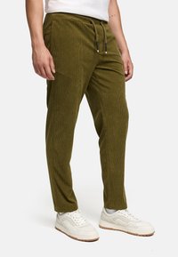 Uomo che indossa pantaloni in corda verde oliva con vita a cordoncino e sneakers bianche, in piedi contro uno sfondo bianco.