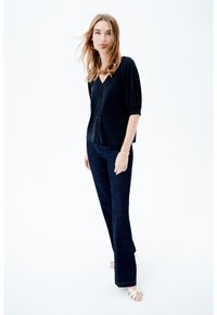 Blusa negra de botones con mangas abullonadas, combinada con pantalones de mezclilla oscuros de pierna acampanada y sandalias planas de color claro. Fondo liso.