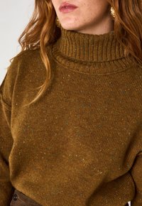 Maglione marrone a maglia grossa con colletto alto a coste, caratterizzato da piccole macchie di blu e giallo su tutto il tessuto. Texture morbida.