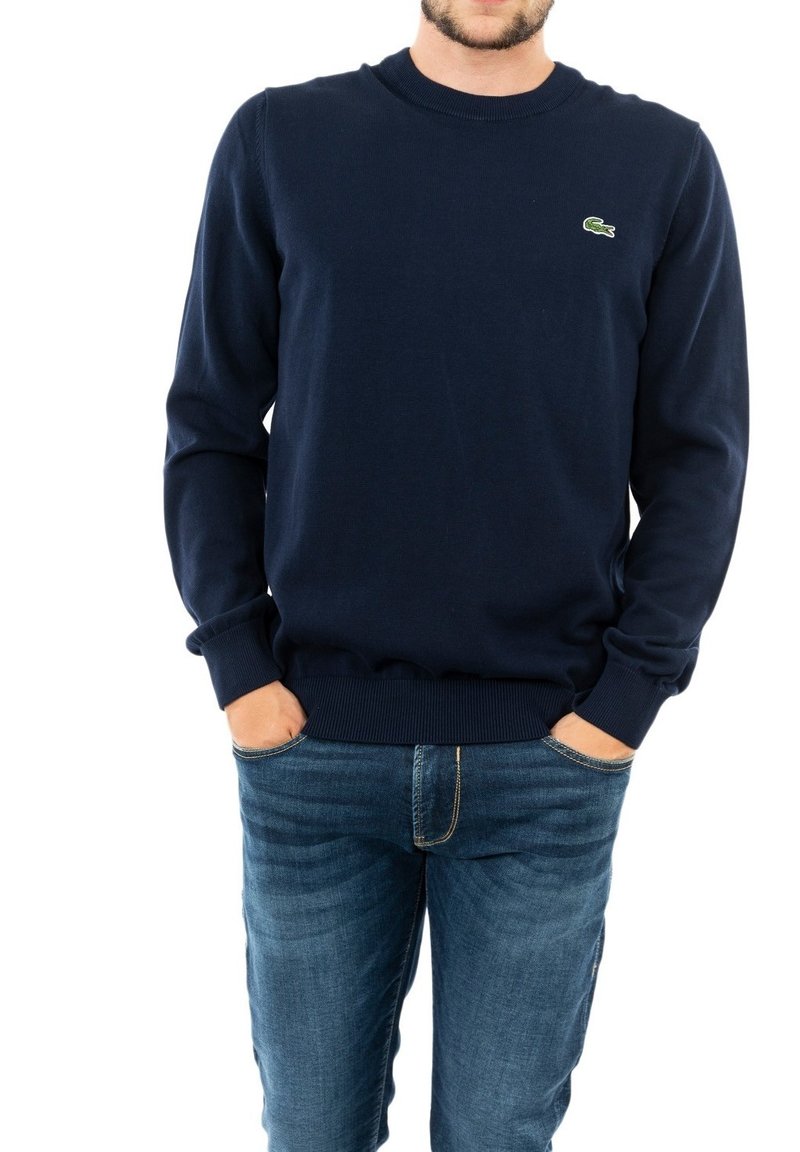 Suéter de cuello redondo azul marino hecho de algodón, con un logo verde de Lacoste. Puños y dobladillo acanalados, combinado con jeans de color azul claro.