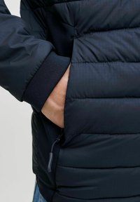 Marineblå pufferjakke med kviltet tekstur, ribbede manchetter og sidelommer. Har en lynlås med træk og en let glans.