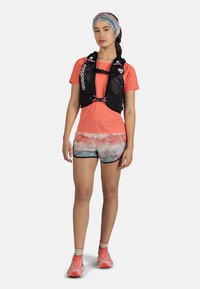 Black hydration vest with logo, bright coral shirt, patterned shorts in pink and blue hues, and pink running shoes with contrasting accents.