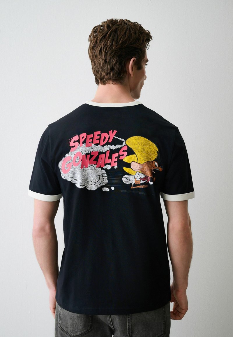 Homme portant un t-shirt noir avec une bordure blanche, avec un dessin animé coloré de Speedy Gonzales et du texte au dos contre un mur uni.