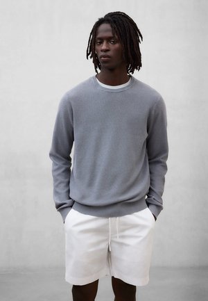 Ung mann med mørke dreadlocks som har på seg en grå langermet genser og hvite shorts, stående med hendene i lommene mot en enkel bakgrunn.