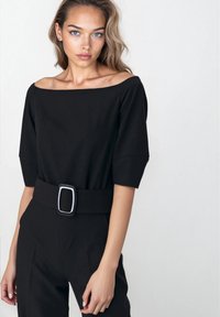 Kobieta o niebieskich oczach i falujących blond włosach, ubrana w czarny top bez ramion oraz spodnie z wysokim stanem z szerokim paskiem.