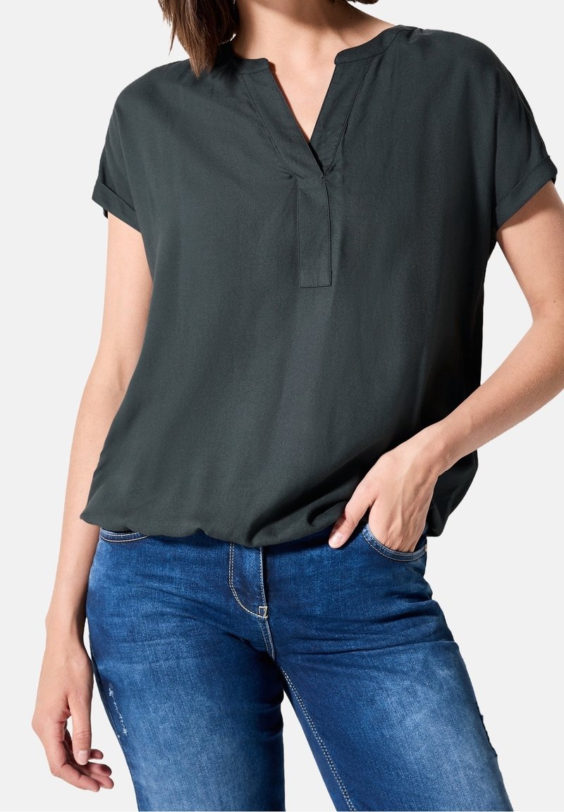 Femme portant un chemisier vert foncé à manches courtes avec un décolleté en V et un jean bleu, une main dans la poche, sur un fond uni.
