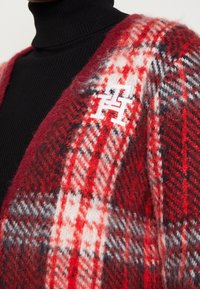 Cardigan à carreaux rouge et noir en tissu doux et texturé. Présente un logo blanc brodé sur le côté gauche. Conception structurée à l'avant ouvert.