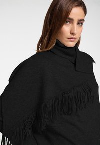 Schwarzer Rollkragenpullover mit einem Fransenakzent an einer Seite, aus strukturierter Strickware und mit asymmetrischem Design.