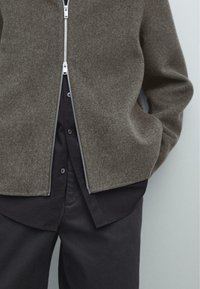 Giacca in lana marrone con zip, dal colletto alto, indossata sopra una camicia nera con bottoni, caratterizzata da una zip liscia e un tessuto strutturato.