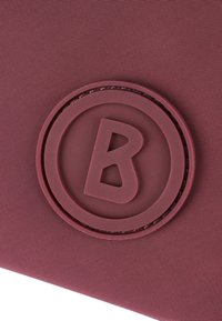 Bordó texturovaná tkanina s kulatým gumovým logem, které má ve středu vyříznutý design písmene 'B'. Kulaté logo je zvýrazněno vyvýšeným okrajem.
