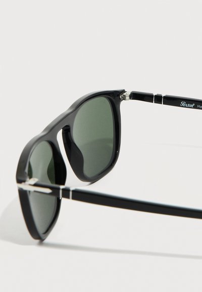 Μαύρα γυαλιά ηλίου με ορθογώνιο σκελετό και σκούρους πράσινους φακούς, διακοσμημένα με ασημένιες λεπτομέρειες στην άρθρωση και μπράτσα με επώνυμα λογότυπα, σε λευκό φόντο.