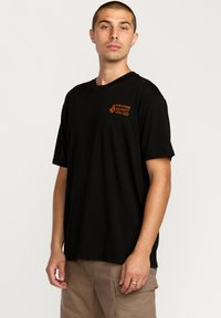 Sort kortærmet t-shirt lavet af bomuld. Den har et orange logo på venstre brystområde. Lige snit med rund halsudskæring.