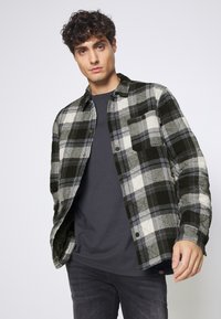 Schwarzes, graues und weißes kaririertes Flanellhemd mit einer Brusttasche, Knopfleiste und weicher Textur, über einem dunklen T-Shirt mit Rundhalsausschnitt.