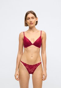 Femme se tenant debout, portant un ensemble coordonné de soutien-gorge et culotte en dentelle rouge, sur un fond clair uni, face à l'objectif avec une expression détendue.