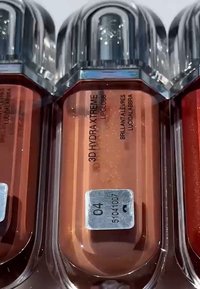 Trois tubes de gloss à lèvres : couleurs ambre, pêche et rouge, avec des bouchons métalliques et des étiquettes indiquant "3D HYDRAXTREME" et les numéros de nuance 04, 51041007.