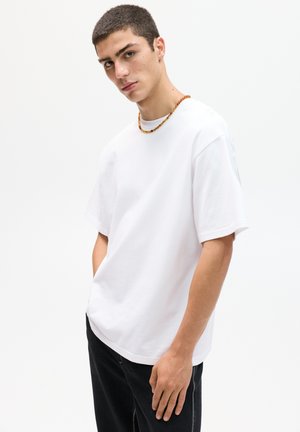 White cotton t-shirt with a relaxed fit, short sleeves, and a round neck. Worn with dark jeans and an amber bead necklace.