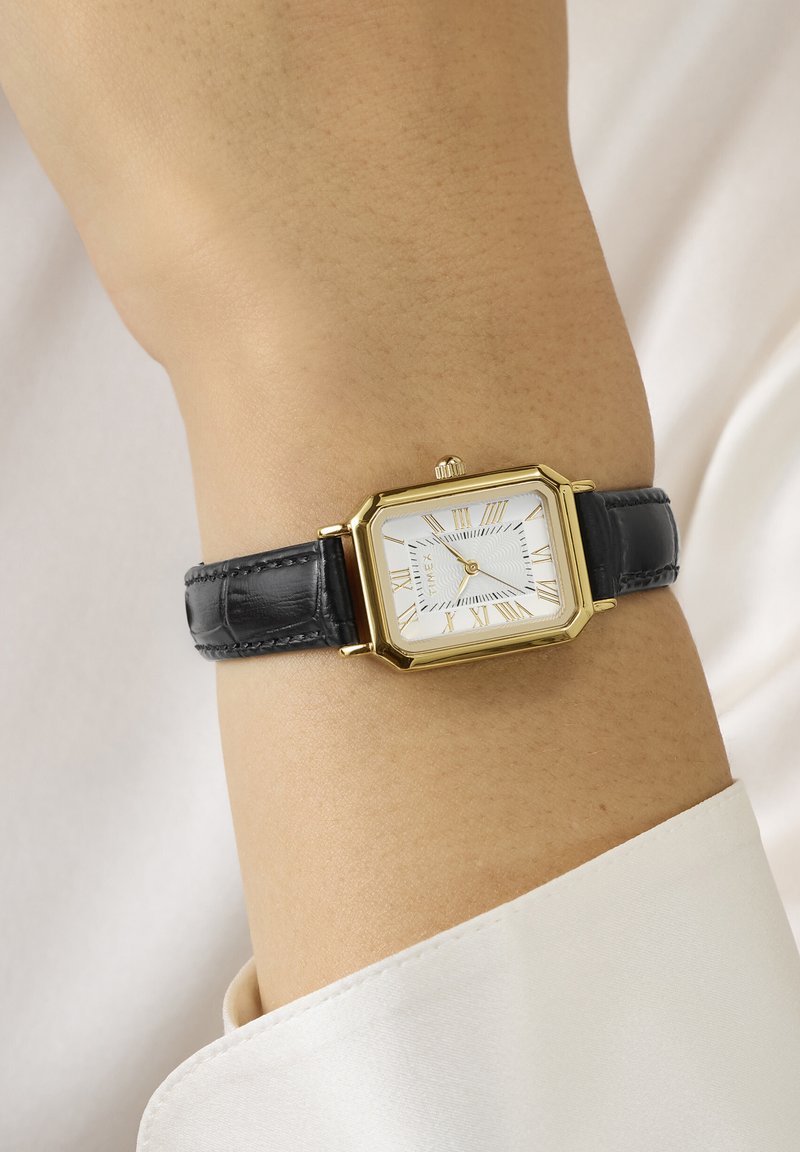 Ορθογώνιο χρυσό ρολόι με λευκό καντράν, ρωμαϊκούς αριθμούς και μαύρο δερμάτινο λουράκι που φοριέται στον καρπό πάνω από την μανσέτα λευκού πουκαμίσου.