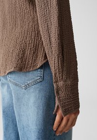 Braunes, texturiertes Langarmhemd mit Knopfmanschette, kombiniert mit hellblauen Jeans mit Rückentaschendesign.}}