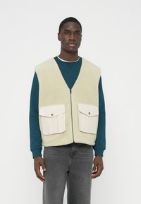 Gilet in pile beige chiaro con scollo a V, cerniera frontale e due tasche con patta color crema. Indossato sopra una maglia a maniche lunghe color teal e pantaloni grigi.