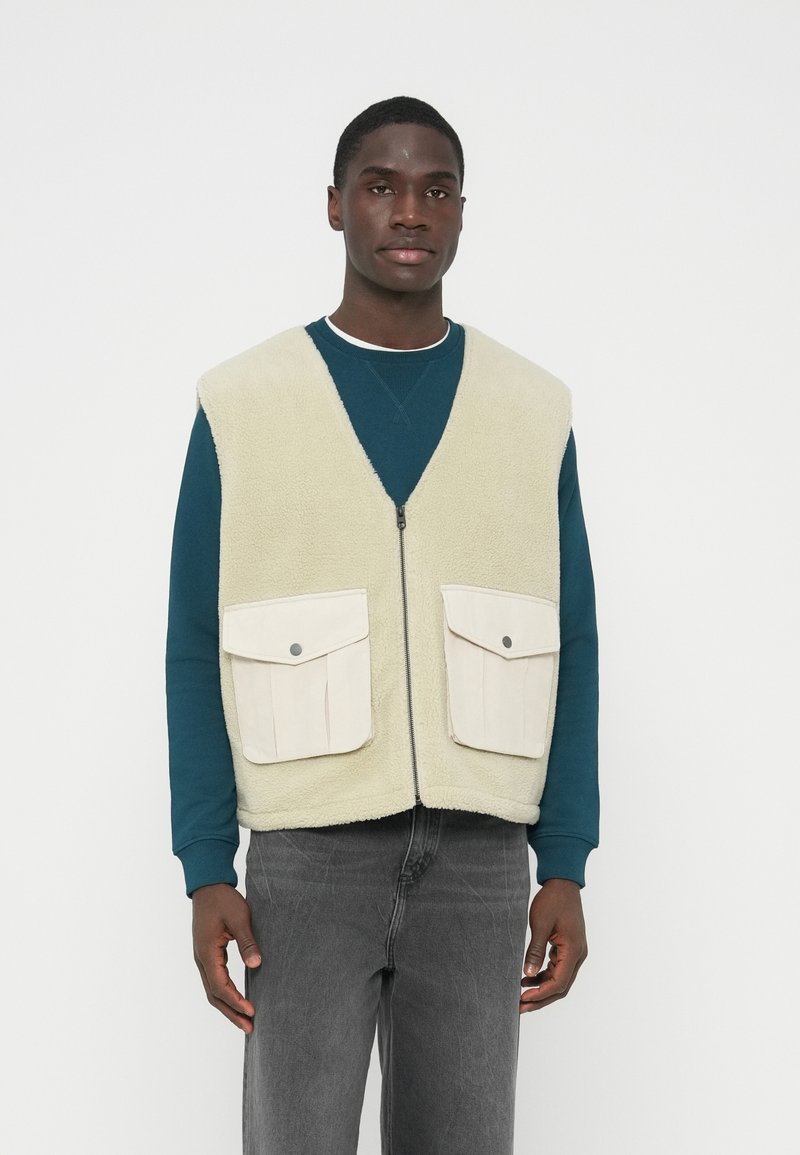 Gilet in pile beige chiaro con scollo a V, cerniera frontale e due tasche con patta color crema. Indossato sopra una maglia a maniche lunghe color teal e pantaloni grigi.