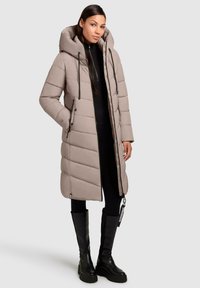 Vrouw in een lange beige gewatteerde pufferjas met capuchon, zwarte leggings en zwarte kniehoge laarzen, tegen een effen achtergrond.