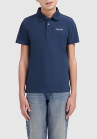 Marineblauwe polo gemaakt van katoen met een kraagdesign, drie knopen en een wit logo op de linkerkant van de borst. Gecombineerd met lichtblauwe jeans.