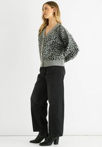 Woman wearing grey leopard print cardigan, loose black jeans, and black heeled ankle boots, standing against a plain white background.