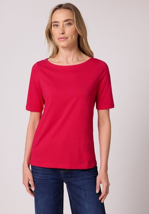 Femme portant un haut rouge vif à manches courtes et un jean bleu foncé, debout devant un fond clair uni.