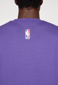 T-shirt de algodão roxo com decote redondo e uma pequena etiqueta com o logótipo da NBA nas costas. O tecido parece suave e tem uma textura lisa.