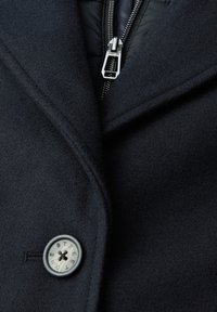 Marineblauwe wolmix jas met een geknipt kraag, zilveren ritssluiting en een ronde knop met "ONE STREET." Gestructureerde stof met een gladde afwerking.