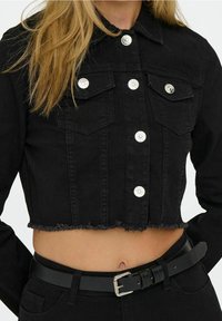 Black cropped denim jacket with silver buttons over bare midriff, paired with black belt and black pants, blonde hair visible.
