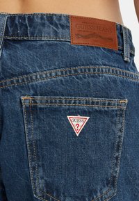 Jeans in denim con un lavaggio blu scuro, dotati di una toppa in pelle dietro e di un logo a triangolo rosso e bianco sulla tasca posteriore.