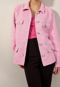 Woman wearing a pink textured jacket with pearl buttons over a darker pink top and black pants against a plain background.