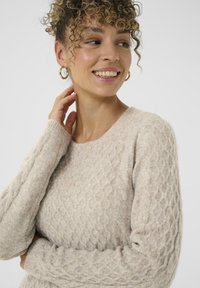 Vrouw met krullend haar in een beige kabelgebreide trui, kijkt opzij en glimlacht, met één hand die haar nek aanraakt.