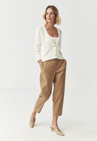 Cardigan bianco a coste con scollo a V e chiusura a bottoni, indossato sopra una maglietta crema, abbinato a pantaloni cropped beige e décolleté neutre a punta.