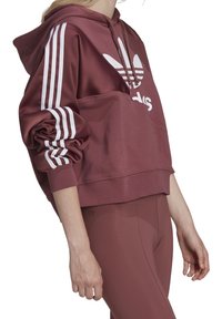 Sudadera corta color burdeos con logo blanco de Adidas, tres rayas blancas en las mangas, tela suave, bolsillo tipo canguro y capucha con cordón.