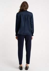 Granatowa satynowa bluzka z marszczonym tyłem, długimi rękawami, mankietami na guziki oraz dopasowanymi eleganckimi spodniami. Noszona z błyszczącymi czarnymi butami.