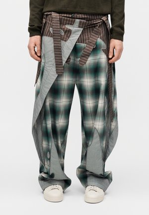 Pantaloni larghi a quadri con toni verdi e grigi, con pannelli di tessuto marrone a righe attaccati, indossati con scarpe sportive bianche e una maglia scura a maniche lunghe.