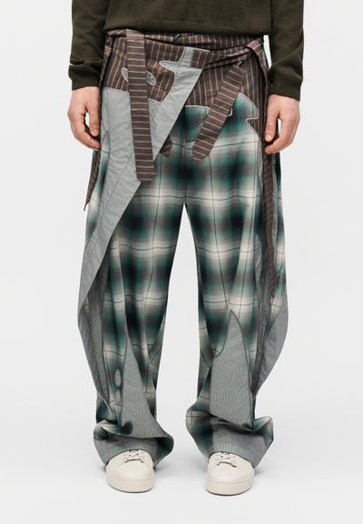 Loose plaid pants with green and gray tones, featuring attached brown striped fabric panels and worn with white sneakers and a dark long-sleeve top.