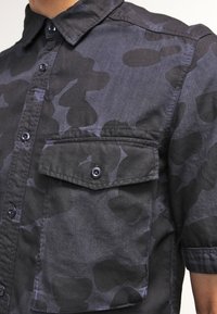 Zbliżenie osoby noszącej krótką koszulę na guziki w ciemnoszarej i czarnej kamuflaży z zapinaną na guziki kieszenią na piersi.