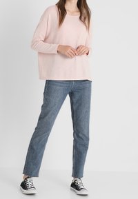 Ljusrosa långärmad topp med avslappnad passform, ihopparad med blå raka jeans och svarta sneakers med vita sulor.