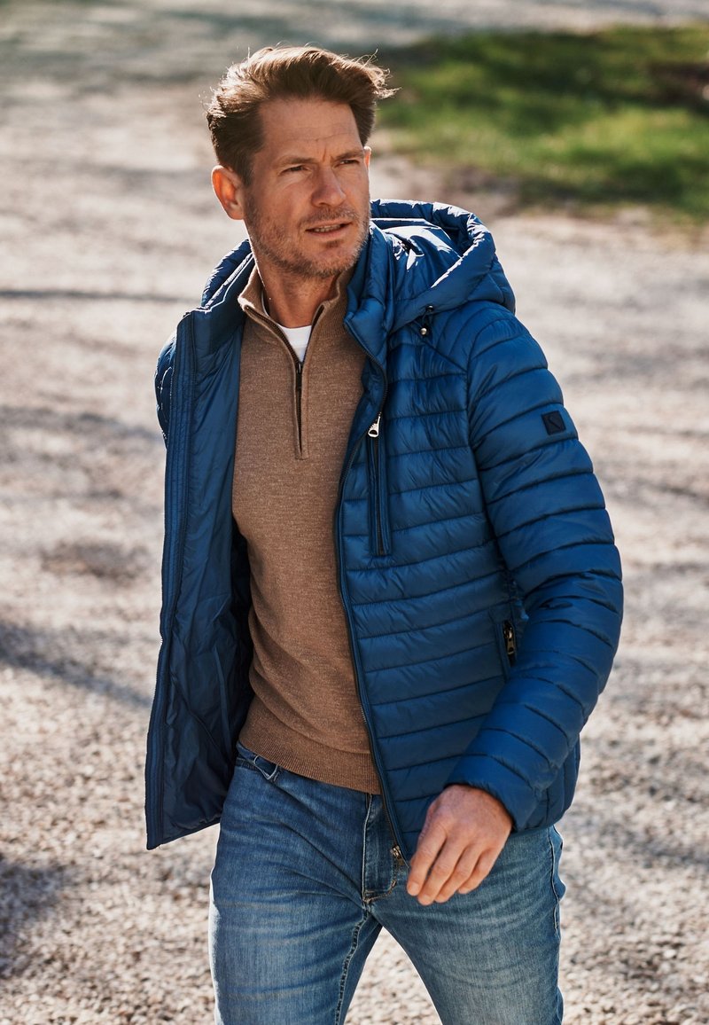 Blauwe pufferjas met een capuchon en ritszakken, gelaagd over een bruine trui, gecombineerd met blauwe spijkerbroek. Getextureerd, gewatteerd materiaal.